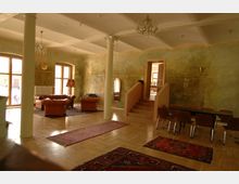 Innenraum eines gemütlichen Wohn- oder Aufenthaltsraums mit pastellgrünen Wänden, großen Fenstern, Säulen und Holzboden. Der Raum ist mit Ledersofas, Teppichen, einem Tisch mit Stühlen und einem Kronleuchter ausgestattet, und eine Treppe führt zu einem angrenzenden Bereich.