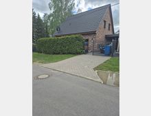 Einfamilienhaus aus Backstein mit steilem, dunkel gedecktem Satteldach und zwei kleinen Fenstern im Giebel. Davor liegen eine gepflasterte Einfahrt mit Metalltor, hohe Hecke und ein kleiner Vorgarten entlang einer Wohnstraße.