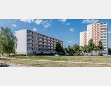 Mehrstöckige Wohnhäuser in einer städtischen Umgebung, links ein flaches Gebäude mit fünf Etagen und rechts ein Hochhaus mit elf Etagen, umgeben von Grünflächen, Bäumen und einem Parkplatz. Der Himmel ist klar mit vereinzelten Wolken.