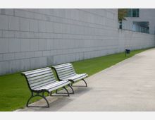 Zwei Holzbänke auf einem Gehweg neben einer Rasenfläche und einer großen weißen Betonwand; im Hintergrund ist ein moderner Gebäudeteil mit Geländer zu sehen.