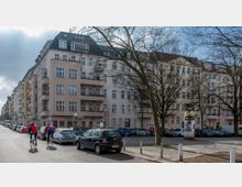 Straßenszene in einem Wohngebiet mit mehrstöckigen Altbauten in hellen Farben und zahlreichen Balkonen. Am Straßenrand parken Autos, während zwei Radfahrer auf der Straße unterwegs sind; im Vordergrund stehen Bäume ohne Laub.