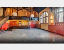 Großer Veranstaltungsraum mit dunklem Holzboden, einer Bar an der linken Wand und großen roten Doppeltüren in der Mitte. Der Raum hat hohe Decken, eine Empore mit Holzgeländer, Backsteinelemente an den Wänden und hohe Fenster mit Rundbögen, durch die Tageslicht einfällt.