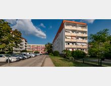 Mehrstöckige Wohngebäude mit Balkonen in einer Wohnsiedlung, umgeben von Bäumen und Grünflächen. Im Vordergrund verläuft ein gepflasterter Gehweg, daneben befinden sich parkende Autos entlang einer Straße.