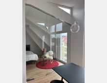 Ein modernes, lichtdurchflutetes Zimmer mit großen Glaswänden, die ein Schlafzimmer mit einem weißen Bett und einem kleinen Sitzbereich abtrennen. Im Vordergrund ein schwarzer Tisch, während im Hintergrund ein roter Teppich, ein Schaukelstuhl mit Fell und Fenster mit Blick auf benachbarte Gebäude zu sehen sind.