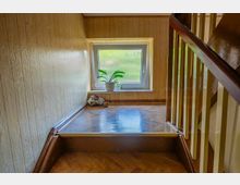 Treppenhaus-Innenraum mit holzvertäfelten Wänden und glänzendem Parkettboden, rechts eine Treppe mit dunklem Handlauf und hellen Geländern. Am Ende des Podests befindet sich ein Fenster mit Milchglas, auf der Fensterbank stehen Topfpflanzen und kleine Dekorationsgegenstände.