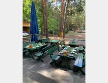 Gedeckte grüne Holzbänke und Tische auf einer gepflasterten Fläche, umgeben von hohen Kiefern in einem Wald. Im Hintergrund sind ein blauer Sonnenschirm, Holzbauelemente und ein Kletterbereich zu erkennen.