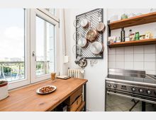 Helle Küche mit großer Fensterfront und Holzarbeitsplatte; an der Wand hängt ein Metallgitter mit Pfannen, daneben offene Holzregale mit Gewürzen und Gläsern. Rechts steht ein Edelstahlherd vor weiß gefliester Rückwand, unter dem Fenster sind Heizkörper und Balkonblick nach draußen zu sehen.