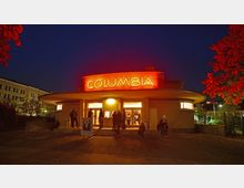 Außenansicht eines Gebäudes im Art-déco-Stil mit geschwungenem Vordach, auf dem in roten, leuchtenden Buchstaben „COLUMBIA“ steht, aufgenommen bei Nacht. Vor dem Eingang stehen oder sitzen mehrere Personen, umgeben von beleuchteten Fenstern und herbstlichem Laub.
