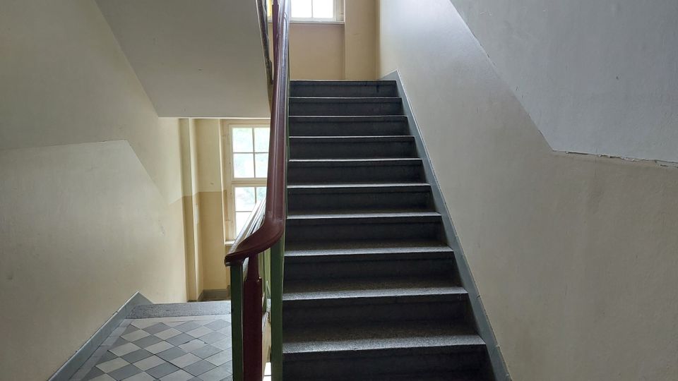Treppenhaus mit grauen Stufen, einem Holzgeländer und hellbeigen Wänden. Im Hintergrund befindet sich ein Fenster, das natürliches Licht einfallen lässt.