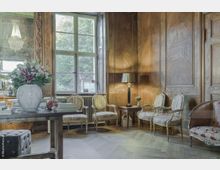 Eleganter Raum mit Holzvertäfelung, antiken Polsterstühlen und einem Tisch mit einer Blumenvase. Große Fenster lassen Tageslicht herein und bieten Blick auf grüne Bäume im Außenbereich.