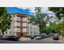 Mehrstöckiges Wohnhaus mit heller Fassade und roten Backsteinelementen an einer Straßenecke, umgeben von Bäumen. Geparkte Autos säumen die Straße, und im Vordergrund stehen ein Verkehrsschild sowie eine Laterne.