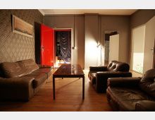 Wohnzimmer mit Holzboden, braunen Ledersesseln und einem braunen Ledersofa. In der Mitte steht ein Holztisch, und im Hintergrund führen farbige Türen in weitere Räume, darunter eine rote Tür und ein abgedunkelter Durchgang.