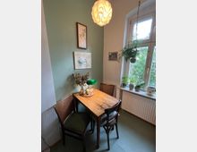 Kleiner Essbereich in einer Wohnung mit einem Holztisch, zwei Korbstühlen und Dekoration wie Kerzen und Trockenblumen. Hinter dem Tisch hängt Wandkunst, und am Fensterbrett stehen Topfpflanzen, während eine Hängelampe die Szene beleuchtet.