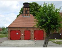 Backsteingebäude mit rotem Ziegeldach, darunter drei rote Holztore und eine Eingangstür. Am Giebel befindet sich ein kleiner Holzturm mit Lamellen, im Vordergrund ein Baum und gepflasterter Gehweg.