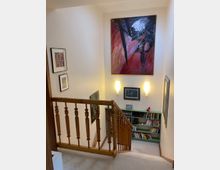 Treppenhaus in einem Wohnhaus mit hellem Teppichboden und einem Geländer aus Holz. An den Wänden hängen mehrere Kunstwerke und ein großes, abstraktes Gemälde. Unterhalb der Treppe befindet sich ein Regal mit Büchern und Dekorationen, beleuchtet von zwei schlichten Wandlampen.