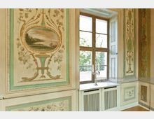 Historischer Innenraum mit pastellfarbenen Wanddekorationsmalereien, darunter Landschaftsmotive und florale Elemente. Großes Fenster mit Holzzargen, das nach außen in eine grüne Gartenlandschaft blickt, und ein silberner Kerzenleuchter steht auf einer weißen Fensterbank über einem Heizkörper.