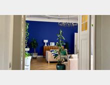 Wohnzimmer mit Holzboden und einer kräftig blauen Wand, darauf ein Sideboard mit Dekoration. Im Raum befinden sich beige Sofas, große Zimmerpflanzen in Töpfen und ein moderner Kronleuchter an der Decke.