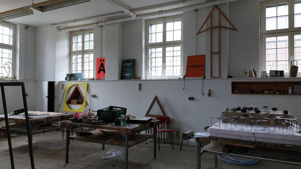 Ein heller Werkraum mit hohen Fenstern und mehreren Holztischen, die mit Werkzeugen, Modellen und Materialien belegt sind. An den Wänden befinden sich künstlerische Plakate, geometrische Rahmen und Regale mit weiteren Utensilien.