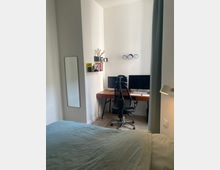 Ein minimalistisches Schlafzimmer mit einem kleinen Arbeitsplatz. Im Vordergrund ein Bett mit grüner Bettwäsche, im Hintergrund ein Schreibtisch mit zwei Monitoren, einem schwarzen Bürostuhl und Regalen an der Wand.