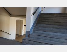 Treppenhaus mit Betonstufen und weiß gestrichenen Wänden, einer Wandhalterung für Handläufe und einer offenen Tür zu einem Nebengang. Ein Stockwerkschild mit der Zahl "3" ist an der Wand angebracht.