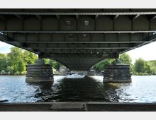 Unteransicht einer stählernen Brücke mit massiven steinernen Pfeilern, die über einen Fluss führt. Im Hintergrund fährt ein kleines Boot unter der Brücke hindurch, umgeben von grünem Laub und einer Uferpromenade.