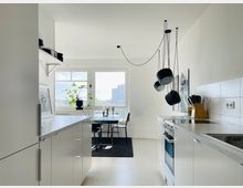 Helle, moderne Küche mit weißer Einrichtung, einer Arbeitsplatte mit Spüle und Herd sowie Hängelampen mit schwarzem Design. Im Hintergrund ein Essbereich mit Tisch und Stühlen vor großen Fenstern, durch die eine städtische Aussicht sichtbar ist.