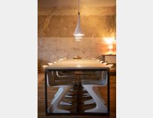 Langer Holztisch mit modernen, weißen Kunststoffstühlen in einem Raum mit rustikaler Betonwand. Eine Pendelleuchte mit drei weißen Lampenschirmen hängt zentral über dem Tisch, im Hintergrund stehen eine Lampe und kleinere Möbelstücke.