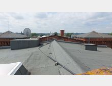 Flachdach eines Gebäudes mit grauer Dachpappe und Kabelinstallationen, umgeben von roten Backsteinwänden. Im Hintergrund sind weitere Gebäudedächer erkennbar, Satellitenschüsseln und eine Landschaft mit Bäumen und blauem Himmel.