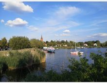 Flussufer mit Schilfbewuchs im Vordergrund, einem Segelboot und einem Ausflugsboot auf dem Wasser. Im Hintergrund sind eine Kirche mit spitzem Turm, eine Windmühle und Häuser in einer kleinen Ortschaft zu sehen, umgeben von Bäumen und unter einem blauen Himmel mit vereinzelten Wolken.