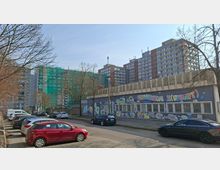 Straßenszene mit parkenden Autos vor einem langgestreckten Flachbau, dessen Fassade mit großflächigem Graffiti und vergitterten Fenstern gestaltet ist. Dahinter stehen mehrere hohe Wohn- oder Bürohochhäuser, teils mit grünem Baugerüst und Netzverkleidung, umgeben von kahlen Bäumen.