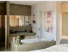 Modernes Badezimmer mit frei stehender Badewanne in Steinoptik, großem Spiegel über einem Waschbecken mit Ablagefläche und dekorativen Elementen wie einer kleinen Skulptur. An der Wand hängen abstrakte Gemälde, und die Einrichtung kombiniert neutrale Farben mit funktionalem Design.