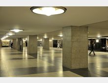 Unterirdische Passage mit großen, rechteckigen Säulen aus grauem Stein, hellen Deckenleuchten und einem Boden aus beigen und grauen Fliesen. Im Hintergrund sind Treppen, Gittergeländer und wenige Passanten zu sehen.