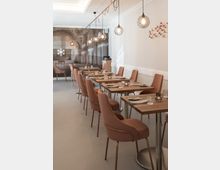 Ein elegantes Restaurant mit modernen Holz-Tischen und gepolsterten Stühlen in Rosa. An der Decke hängen runde, gläserne Pendelleuchten, und eine Wand ist mit einer Fotografie eines historischen Gebäudes dekoriert, daneben eine Wanddekoration mit kleinen Vogelmotiven.