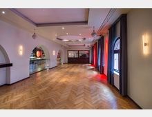 Geräumiger Innenraum mit Parkettboden, weißen Wänden und abgerundeten Deckenleuchten. Links befinden sich mehrere Rundbögen mit Blick auf eine Bar, rechts Fenster mit langen Vorhängen, teils in rotem Licht beleuchtet.