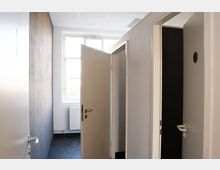 Schmaler Innenflur mit hellen Wänden und abgehängter Rasterdecke, am Ende ein großes Fenster über einem Heizkörper. Mehrere Türen stehen offen und führen in angrenzende, teils dunkle Räume.