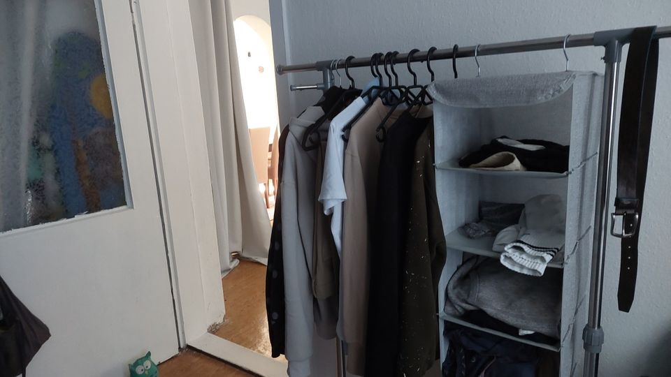 Ein offener Kleiderbereich in einer Wohnung mit einer Metall-Kleiderstange, an der mehrere Kleidungsstücke auf Bügeln hängen, sowie einem hängenden Stoffregal mit gefalteter Kleidung. Links ist eine Tür mit Milchglas zu sehen, daneben ein schmaler Flur mit Holzboden.