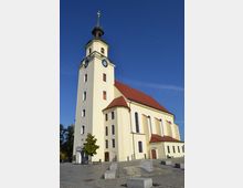Gelbe Kirche mit roten Dachziegeln und einem hohen Turm mit Uhr und kupfernem Spitzdach, gelegen an einem gepflasterten Platz mit kleinen modernen Brunnen. Der Himmel ist klar und blau, und einige Bäume stehen am Rand des Platzes.