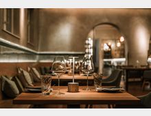 Innenraum eines modern eingerichteten Restaurants mit warmem, gedämpftem Licht, Holztischen und gepolsterten Sitzbänken an der Wand. Im Vordergrund ein gedeckter Tisch mit Weingläsern, Wassergläsern und kleiner Tischlampe; im Hintergrund ein Rundbogen-Durchgang zu weiteren Sitzbereichen.
