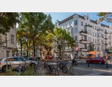 Eine Straßenecke in einem Wohnviertel mit historischen, mehrstöckigen Gebäuden in heller Fassadengestaltung und verzierten Balkonen. Im Vordergrund befinden sich geparkte Fahrräder, Bäume spenden Schatten, und Autos sowie ein Lastwagen sind auf der Straße zu sehen.