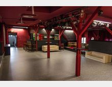 Eine Veranstaltungs- oder Clubräumlichkeit mit roten Wänden und schwarzen Deckenbalken. Der Raum enthält eine Bar mit Barhockern, verschiedene Sitzgruppen mit Sofas, und Bühnenbeleuchtung an der Decke.