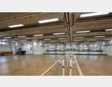 Großer Tanz- oder Übungsraum mit Holzboden, Spiegelwand und fest installierten Ballettstangen. Die Decke ist mit Leuchtstofflampen und sichtbaren Rohren ausgestattet, während große Fenster Tageslicht hereinlassen.