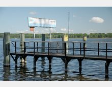 Steg mit Geländer am Wasser, beschildert mit „Station Glienicker Brücke - 2“; im Hintergrund ein Fluss oder See mit Ufer und Bäumen unter klarem Himmel.