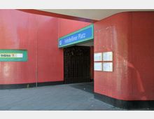 Eingang zur U-Bahn-Station Fehrbelliner Platz mit einer blau-grünen Beschilderung über der Tür, umgeben von roten Fliesen. Daneben befinden sich Aushänge mit Fahrplänen und ein Imbiss-Schild in einer grünen Einfassung.