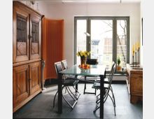 Esszimmer mit modernem und traditionellem Stil: Glas-Esstisch mit schwarzen Metallstühlen, massive Holzschränke, ein orangefarbener Heizkörper, ein großes Fenster mit Blick auf eine Wohngegend und Dekorationen wie gelbe Kerzen und Blumen.