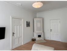 Wohnzimmer mit weißen Wänden und Holzboden, ausgestattet mit einem klassischen weißen Kachelofen in der Ecke, einer Couch im Vordergrund und zwei weißen Innentüren mit klassischen Verzierungen. Eine Lampe mit Stoffschirm hängt an der Decke, und ein gerahmtes Bild sowie ein Fernseher sind an der Wand angebracht.