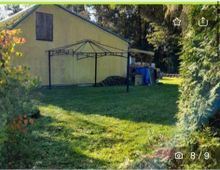 Gelber Schuppen mit geneigtem Dach, umgeben von einer grasbewachsenen Fläche und Bäumen im Hintergrund. Im Vordergrund ist ein Metallrahmen einer Pergola zu sehen, daneben gestapelte Holzscheite.