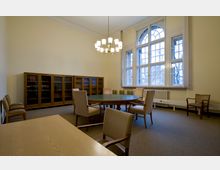 Ein Konferenzraum mit einem runden Holztisch und gepolsterten Stühlen, ergänzt durch eine große Fensterfront mit Rundbogen und Sprossenfenstern. An der Rückwand steht eine Bücherwand mit Glastüren, und die Decke ist mit einem zentralen Kronleuchter beleuchtet.