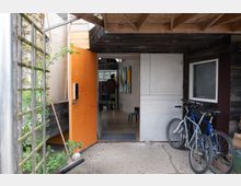Ein Eingang zu einem Gebäude mit einer geöffneten, orangefarbenen Tür. Außen stehen zwei Fahrräder angelehnt, und neben der Tür befinden sich Holzwände und eine Rankhilfe mit grünen Pflanzen. Im Inneren sind Möbel und eine person im Hintergrund zu sehen.