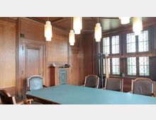 Holzvertäfelter Konferenzraum mit einem großen rechteckigen Tisch in der Mitte, umgeben von Stühlen mit Armlehnen. An der Wand hängen Tafeln mit eingravierten Namen, und ein großes bleiverglastes Fenster lässt Tageslicht herein; moderne Hängelampen hängen von der Decke.