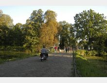 Pflasterstraße mit einem Geländer, die über eine schmale Brücke führt; links und rechts von der Brücke befinden sich kleinere Wasserflächen, umgeben von dichtem Grün. Auf der Brücke fahren ein Motorrollerfahrer und mehrere Fußgänger, die in Richtung eines Eingangs mit hohen Gittertoren gehen.